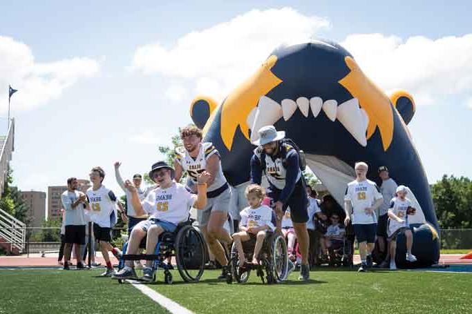 UNC football student-athletes running onto the field, accompanied by disabled children during the No Limits Camp, fostering inclusivity and joy.